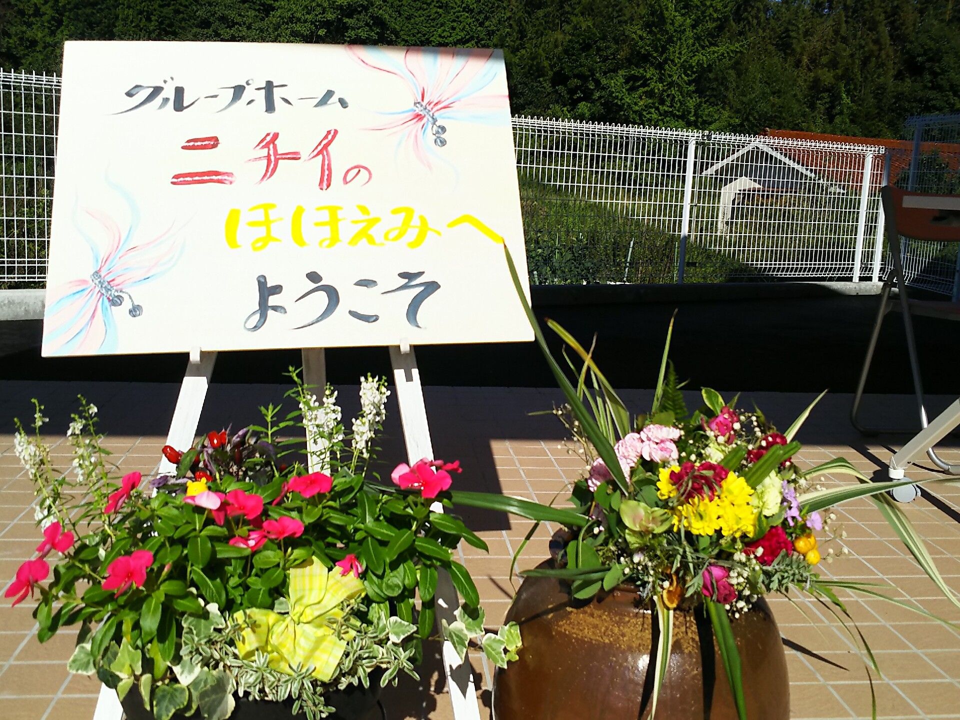 手書きのウエルカムボードがあり、その周りにはカラフルな花の寄せ植えが複数置かれている。