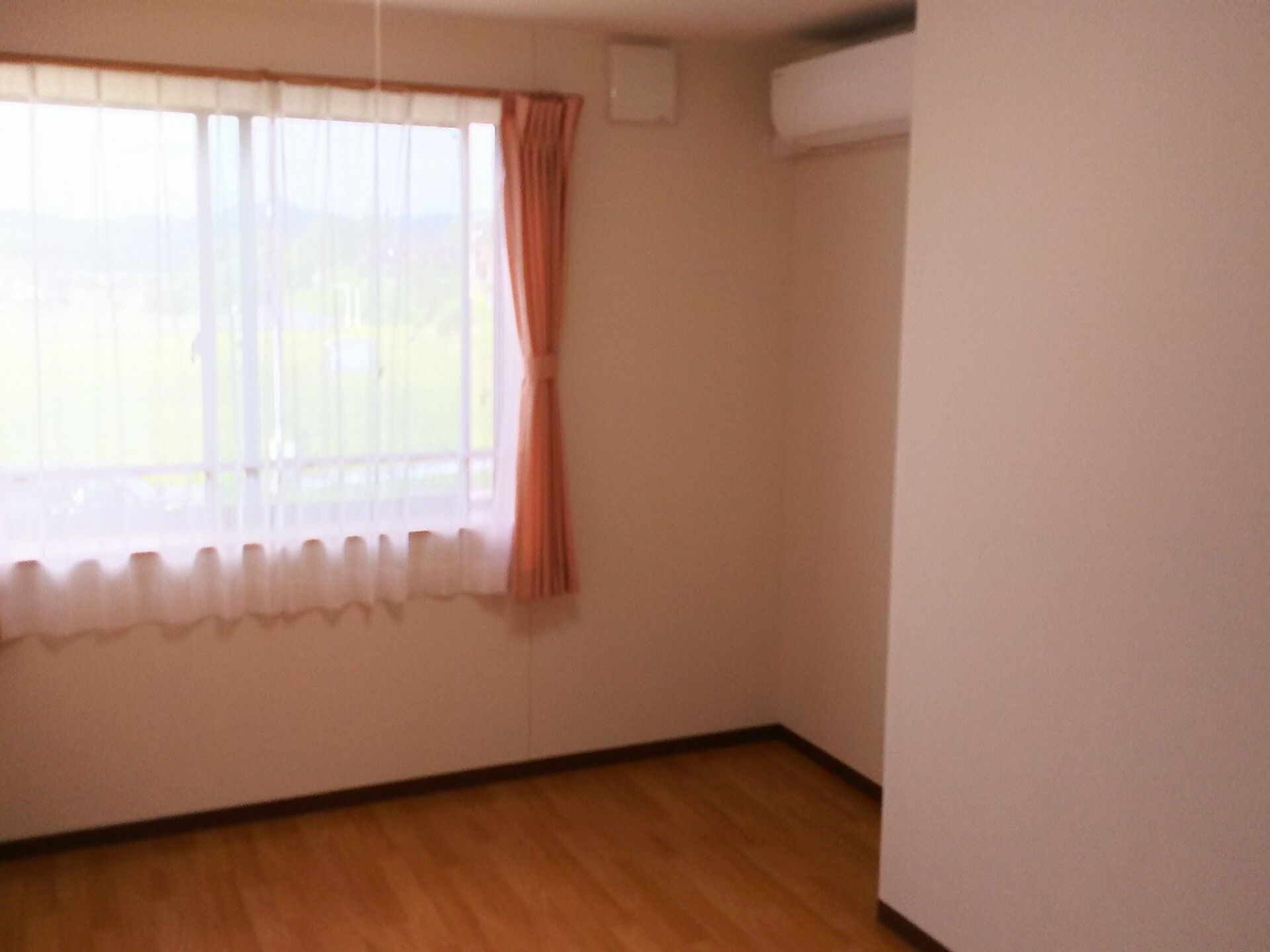 レースカーテンとカーテンが付いた窓があるフローリングの部屋の壁にはエアコンが設置されている。
