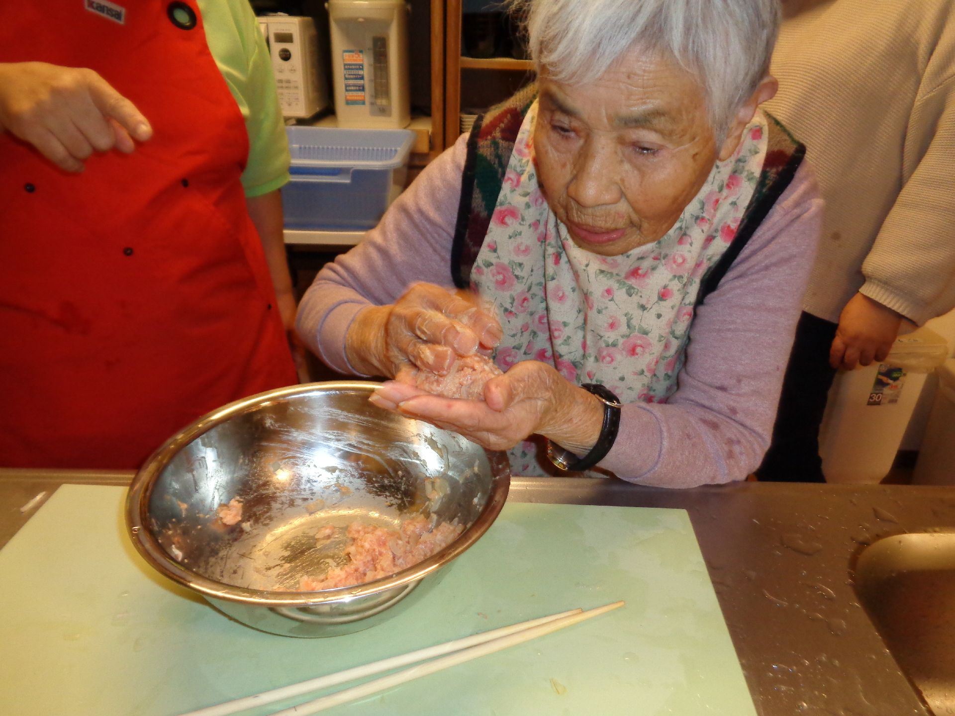 花柄のエプロンを付けた高齢女性が真剣な表情でハンバーグをこねている様子。