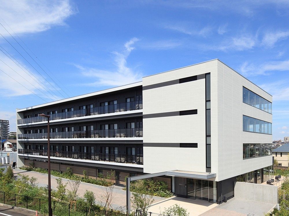 ４階建ての建物の１番下はブラウン、その上のフロアは白い外観で、建物の周りには緑が植えられている。