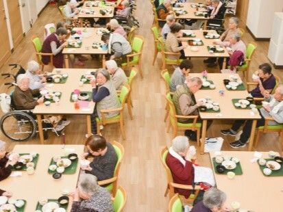 ライトグリーンの椅子がセットされた木製のテーブルが複数置かれた食堂に集まり、入居者が食事をしている。