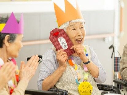 カラー画用紙でできた王冠を頭にのせ、手作りのメダルを首にかけ、大きな手作りのお守りを持って微笑む高齢女性。
