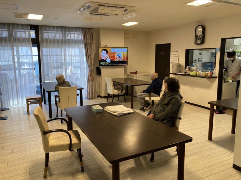 食堂のダイニングテーブルには入居者が数名座っていて、奥にあるキッチンでは作業をしているスタッフがいる。