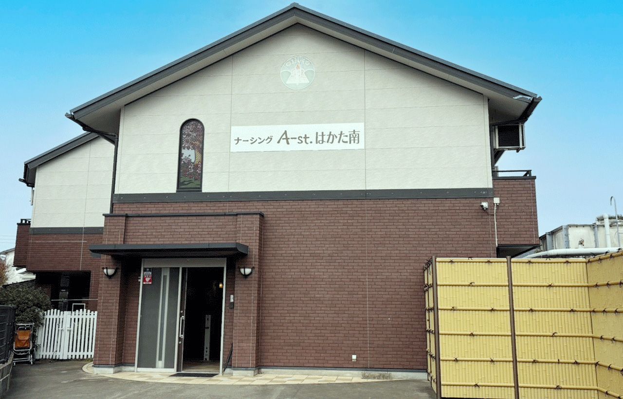 ブラウンとベージュの外壁の2階建ての建物の玄関には屋根があり、外壁にはホーム名が書かれている。