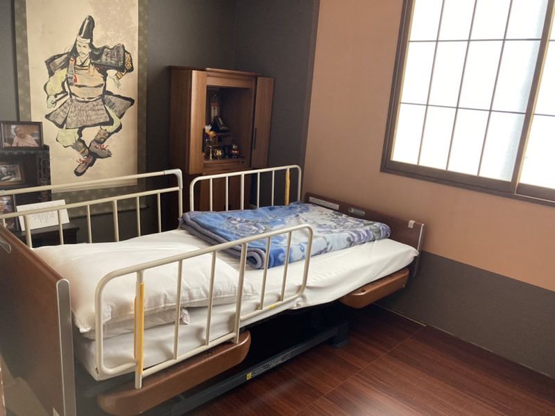 寝具がセットされた介護ベッドがあり、横には侍が描かれた掛け軸があり、仏壇も飾られている。