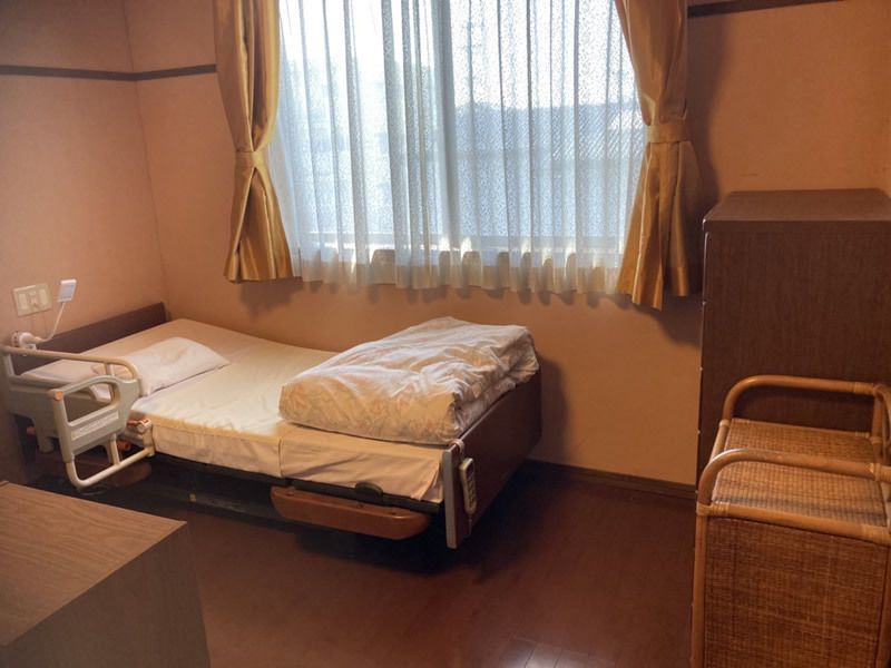 レースカーテンとカーテンが付いた大きな窓がある部屋には寝具付きのベッドが置かれ、家具もある。
