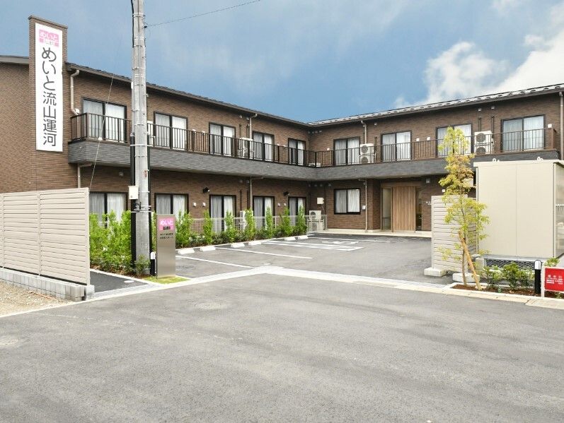 エル字型の建物はブラウン系で敷地内には駐車場があり、草木も植えられている。
