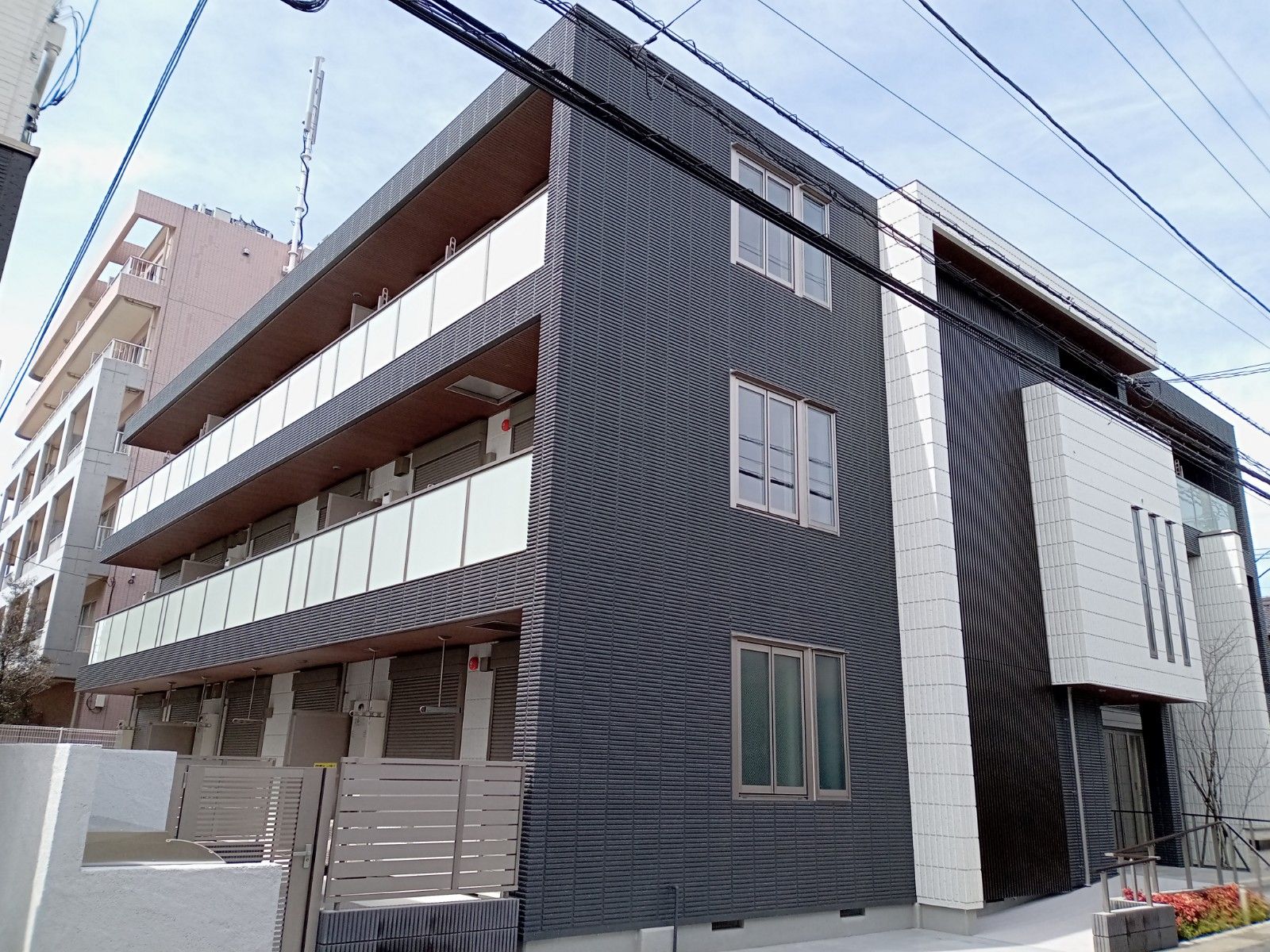 3階建ての建物はダークグレーでまとめられ、各フロアにベランダがあり、奥には建物がある。