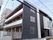 3階建ての建物はダークグレーでまとめられ、各フロアにベランダがあり、奥には建物がある。