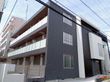 3階建ての建物はダークグレーでまとめられ、各フロアにベランダがあり、奥には建物がある。