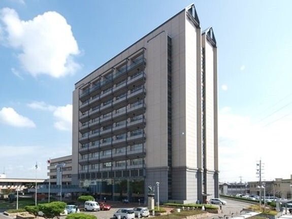 グレー系の大きな建物の周りには草木が植えられ、駐車場には車が止まっている。