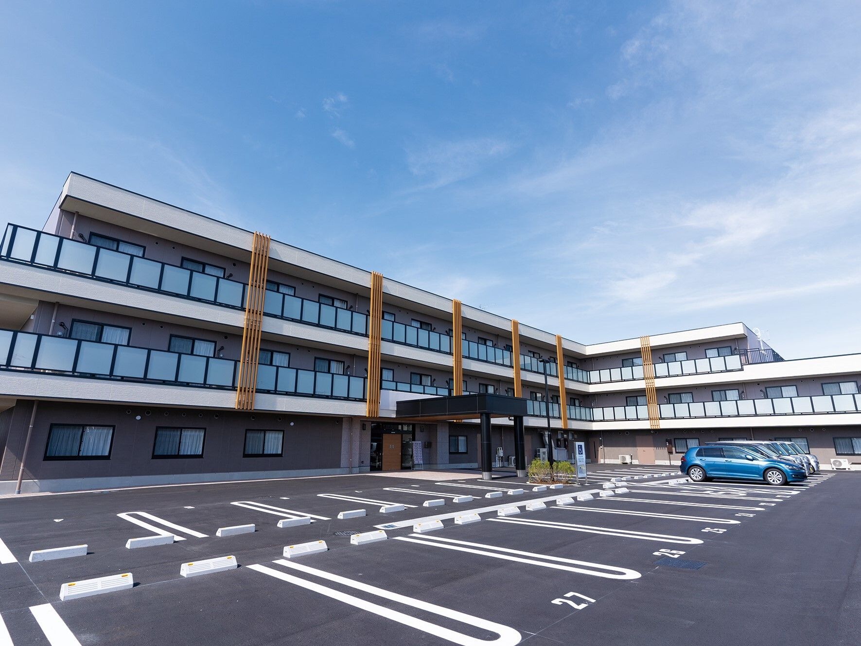 エル字型の3階建ての建物は日当たりのいい場所に建っていて、広い駐車場があり、車が止まっている。