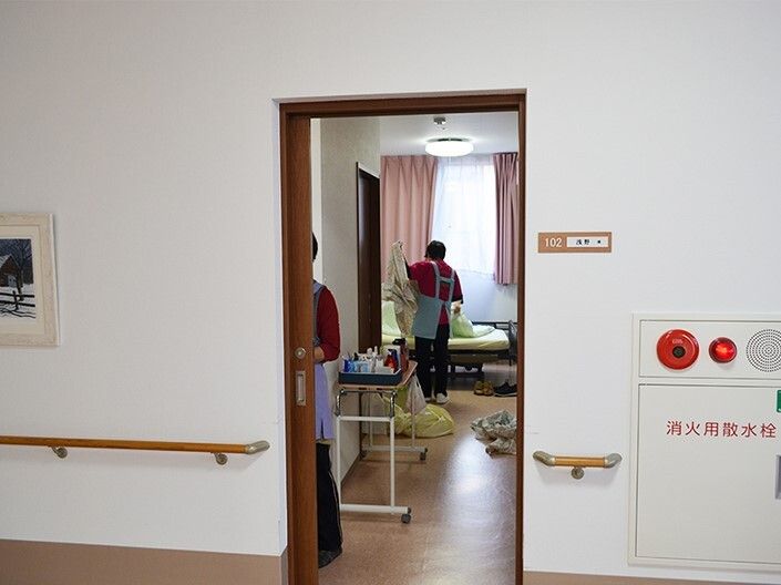 ２名のスタッフが居室内で作業をしている。1名は奥の介護ベッドのシーツを交換している。