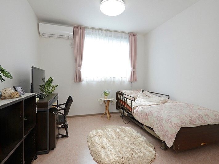 レースカーテンとカーテンが付いた窓がある部屋には寝具付きのベッドやテレビなどが置かれている。