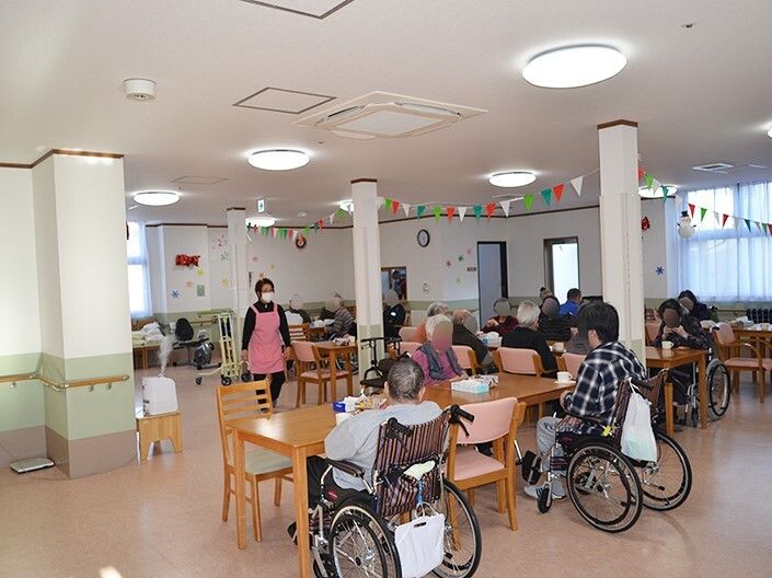 エプロンを付けた女性スタッフが歩いている食堂にはダイニングテーブルが複数あり、入居者が複数名席についている。