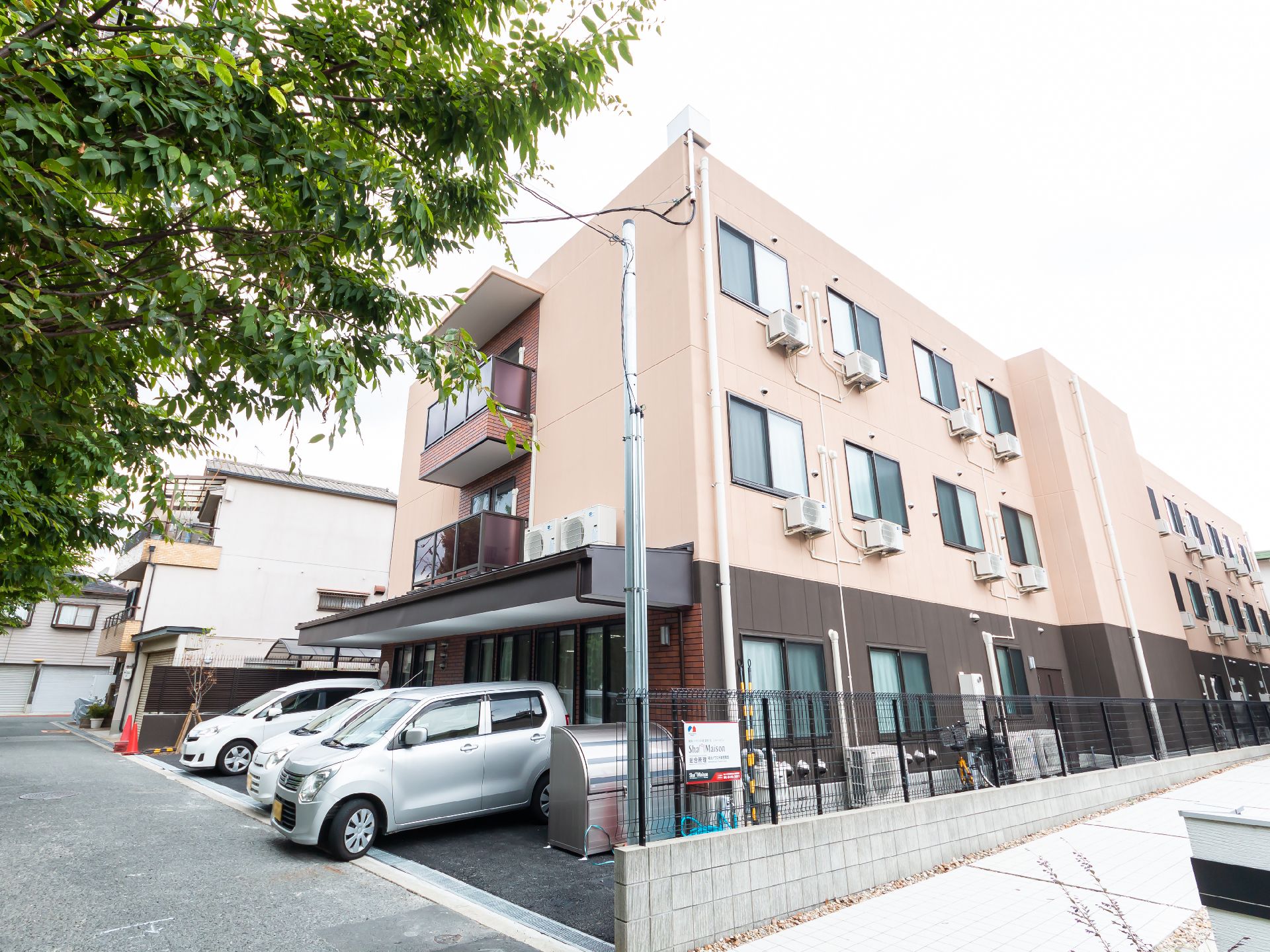 側面の駐車場に車が止まっているベージュの外観で３階建ての建物