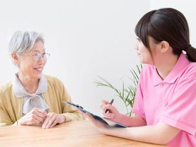眼鏡をかけた高齢女性とピンクのポロシャツを着た女性スタッフがテーブルを囲んで話している。