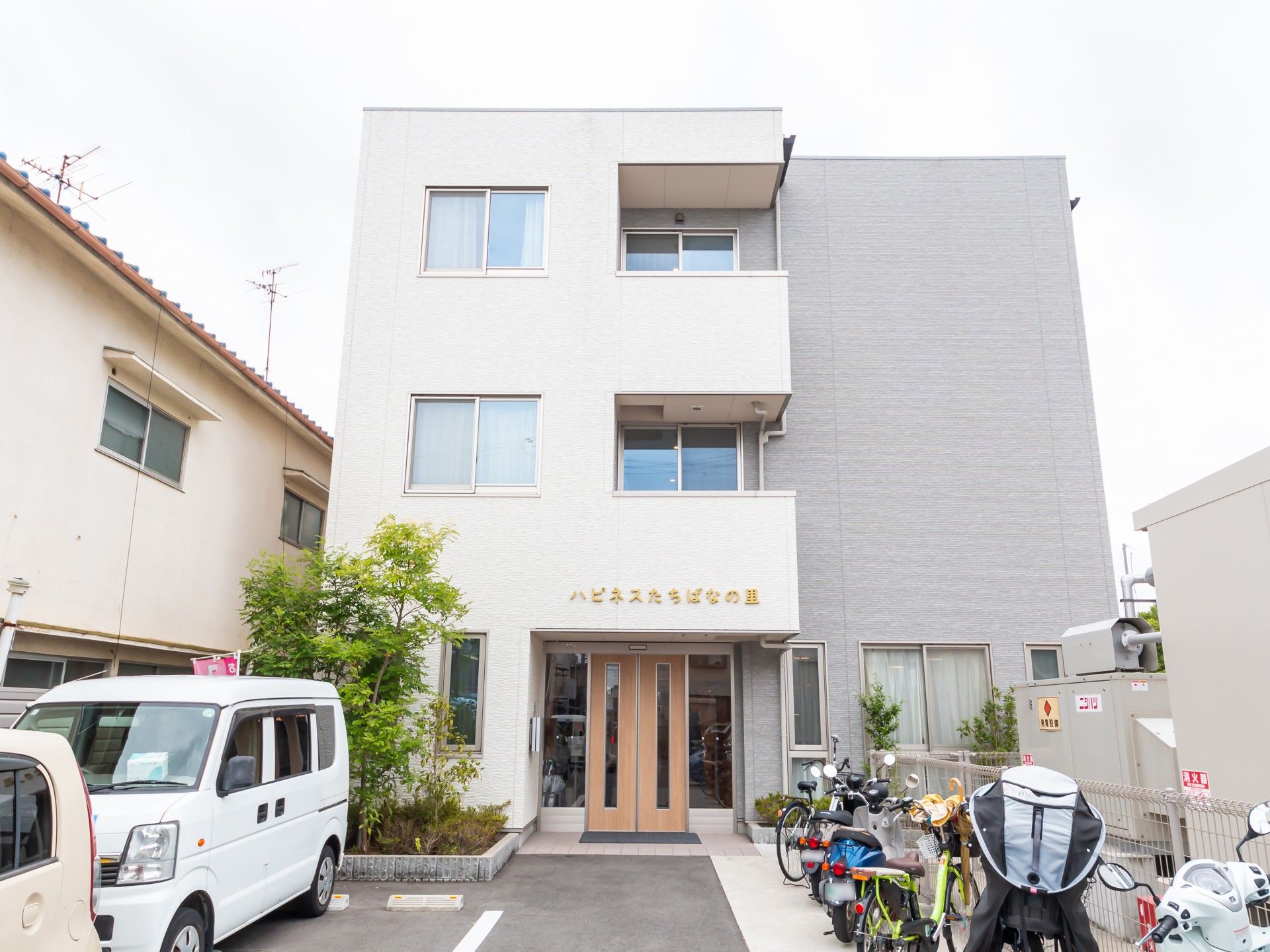 3階建ての建物は白とグレーで敷地内には駐車場もある。外壁にはホーム名が書かれ、草木も植えられている。