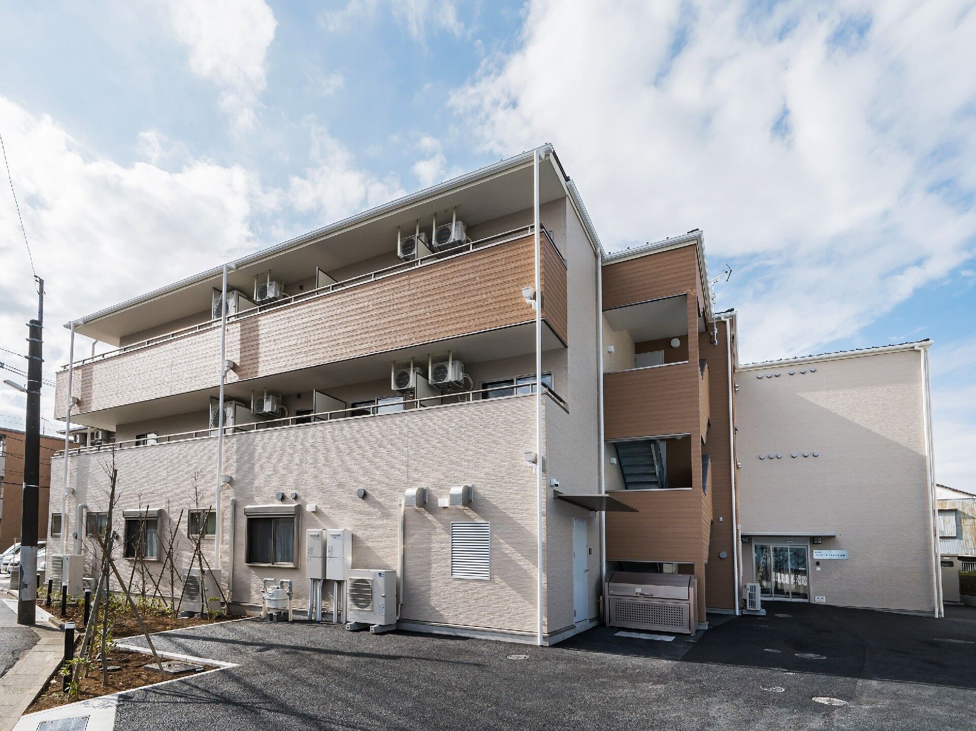 角フロアにベランダが付いたブラウン系の３階建ての建物の前には駐車場がある。