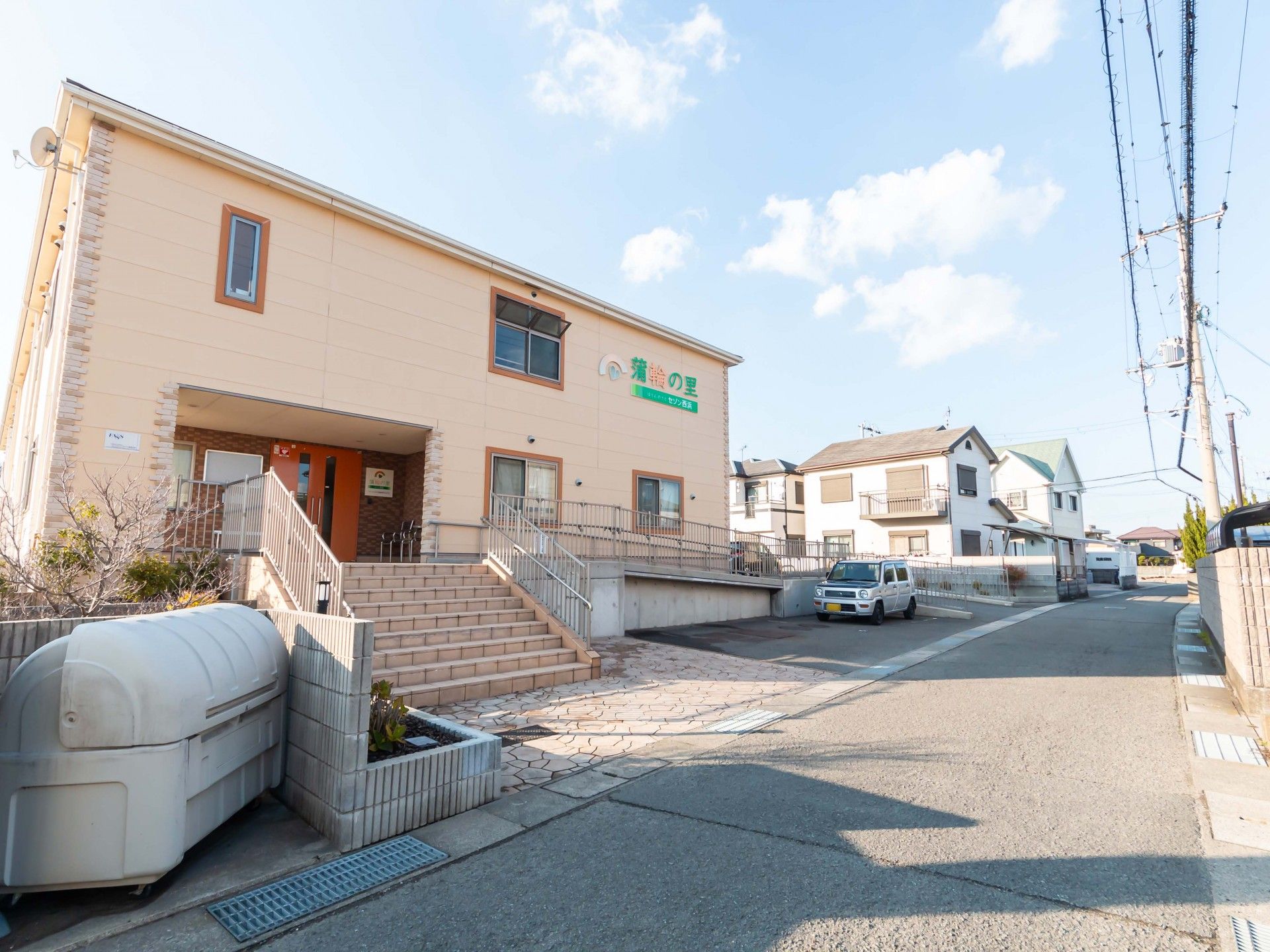 車いすの高齢女性に寄り添って緑の多い屋外で笑顔で過ごしているエプロンを付けた男性スタッフ。