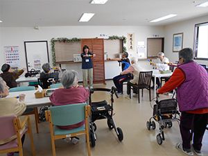 スタッフが前に出てお手本を見せ、スタッフを見習って体操をしている高齢者たち。椅子に座っている。