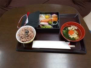 右側には人参が花形に切られた吸い物があり、左側には赤飯があり、中央にはお節風の料理が詰められたお重がある。