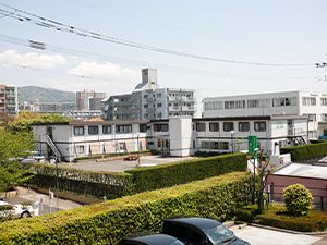 白い2階建ての建物が建つ広い敷地内には駐車場があり、周りにもたくさんの建物が建っている。