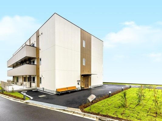 3階建ての建物はグレー系でたくさんの木も植えられている。各フロアにベランダがあり、空は青い。