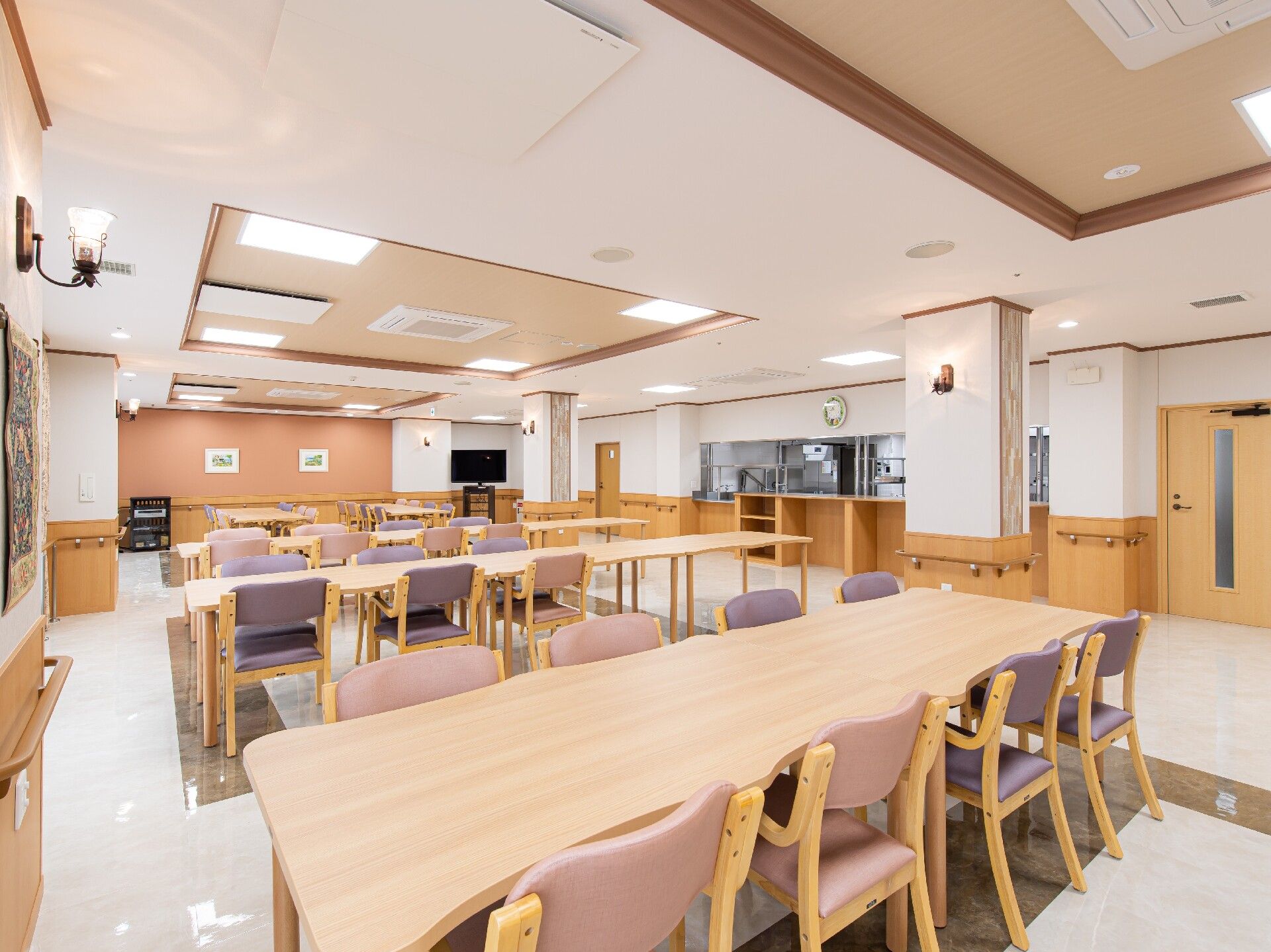 大きなダイニングテーブルセットが複数置かれ明るい雰囲気の食堂。テレビもあり、厨房がすぐそばにある。