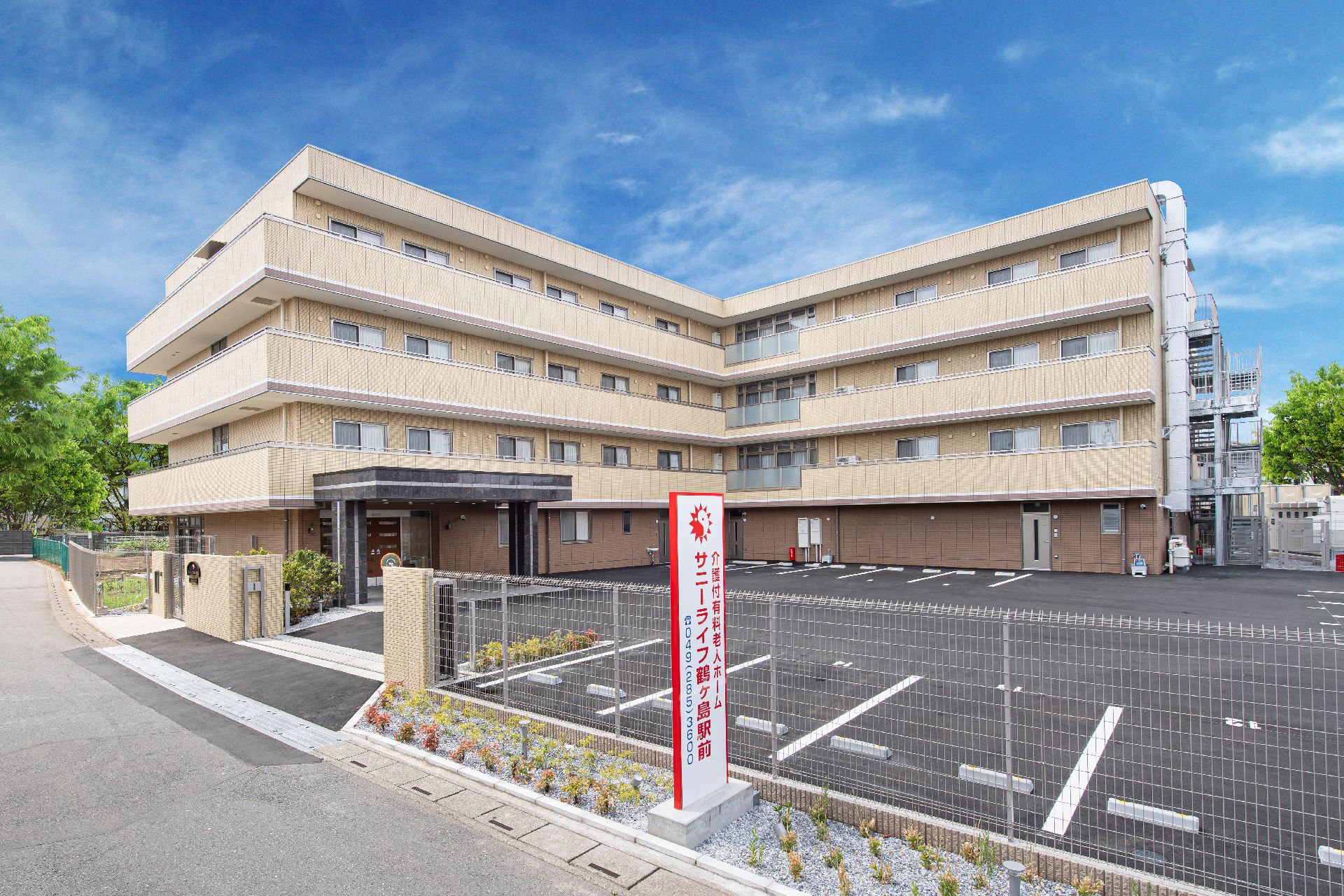 エル字型の建物はベージュ系の外壁で敷地内には広い駐車場がある。4階建ての建物の各フロアにベランダが付いている。