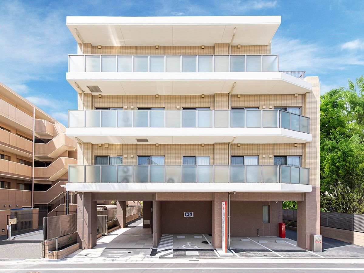 白を基調とした4階建ての建物の１階部分には駐車場があり、周りには緑も残っている。