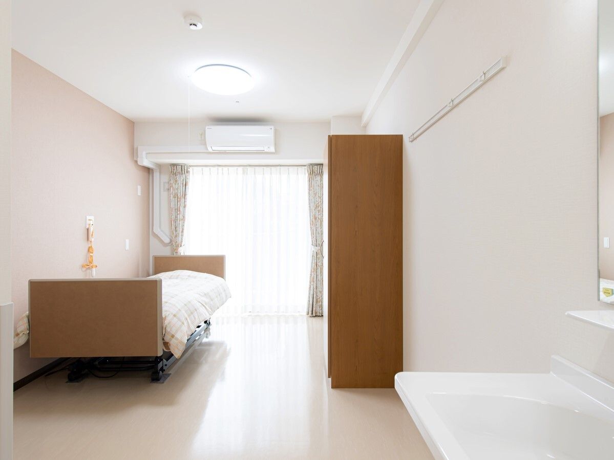 レースカーテンとカーテンが付いている窓がある部屋には寝具付きのベッドがあり、ベッドサイドには緊急通報装置がある。