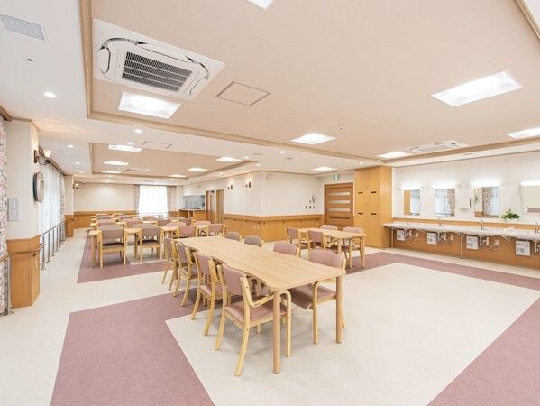 木製のダイニングテーブルセットが複数置かれ、右側には車いす対応の洗面所が並んでいる食堂。