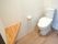 シンプルなデザインのトイレ。壁にはトイレットペーパーホルダーが設置され、角には三角の棚がある。