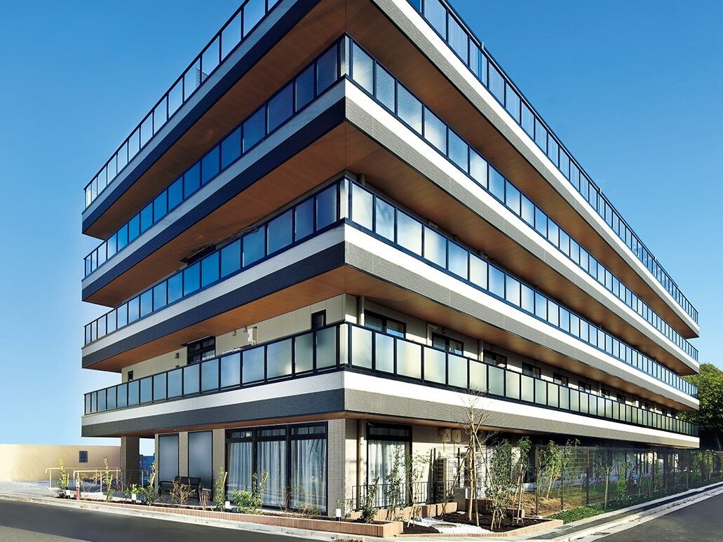グレー系の大きな建物の各フロアにベランダがあり、建物の周辺には木が植えられている。