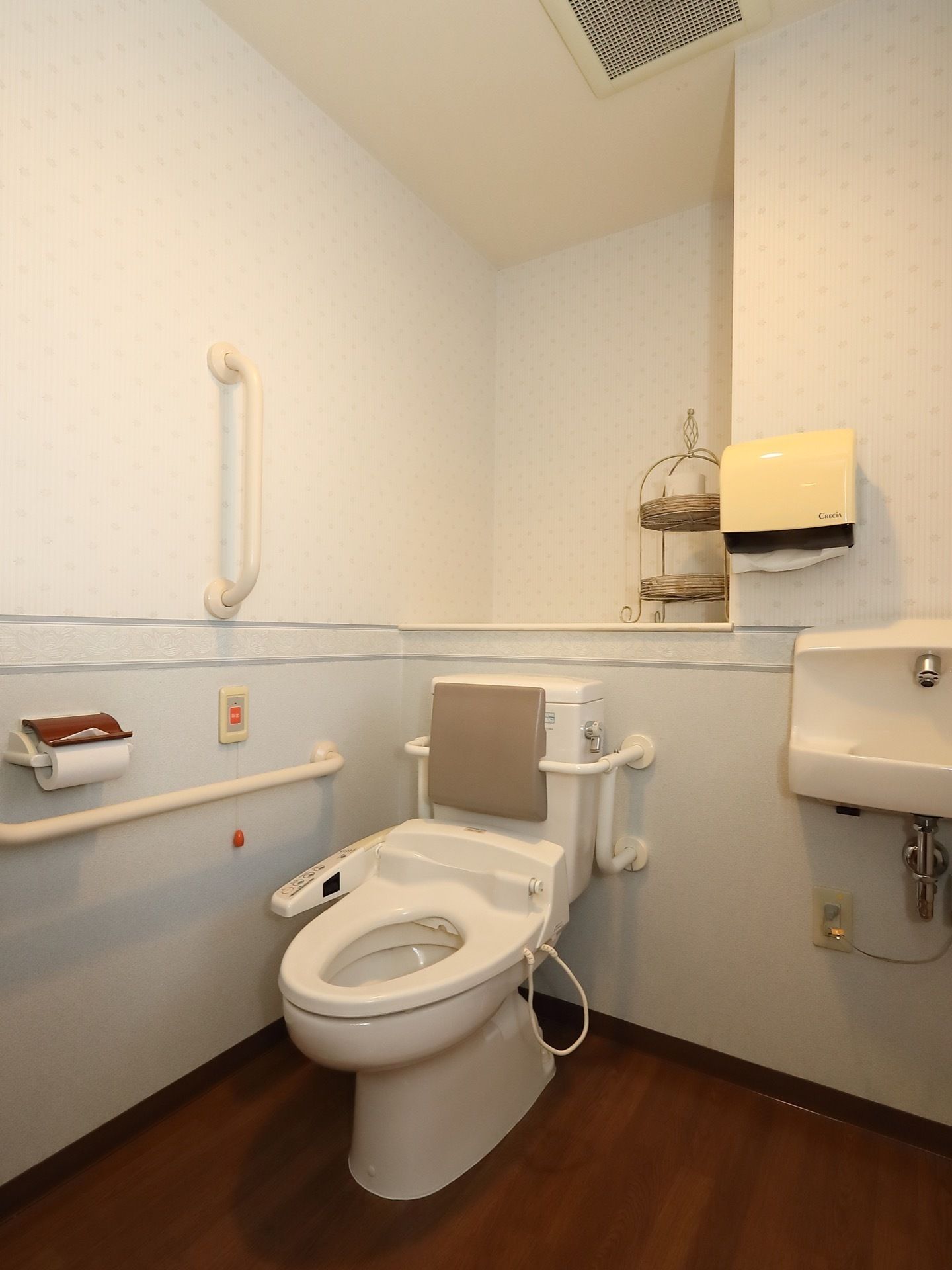 ゆとりある広々としたトイレで、手洗い場が備えつけられている。温水洗浄機能付きの便座には背もたれが設置されている。