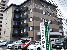 ブラウン系の５階建ての建物の周りにも背の高い建物があり、敷地内には駐車場がある。