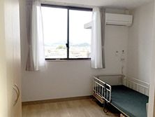 介護ベッドやエアコンが付いたフローリングの部屋にはレースカーテンとカーテンが付いた窓がある。