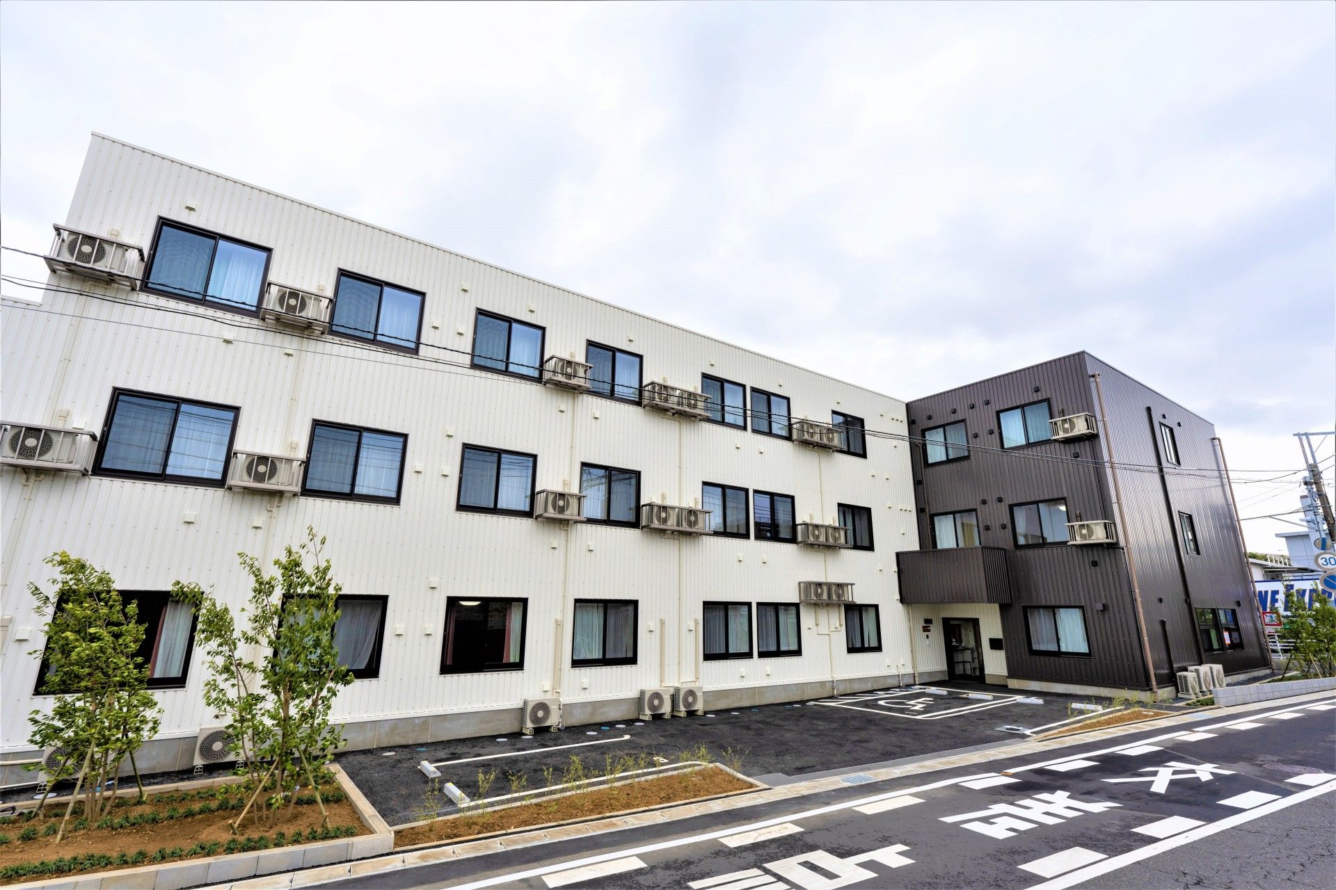 3階建ての建物は白とダークグレーの外壁で建物の周りには背の高さの異なる草木が植えられている。