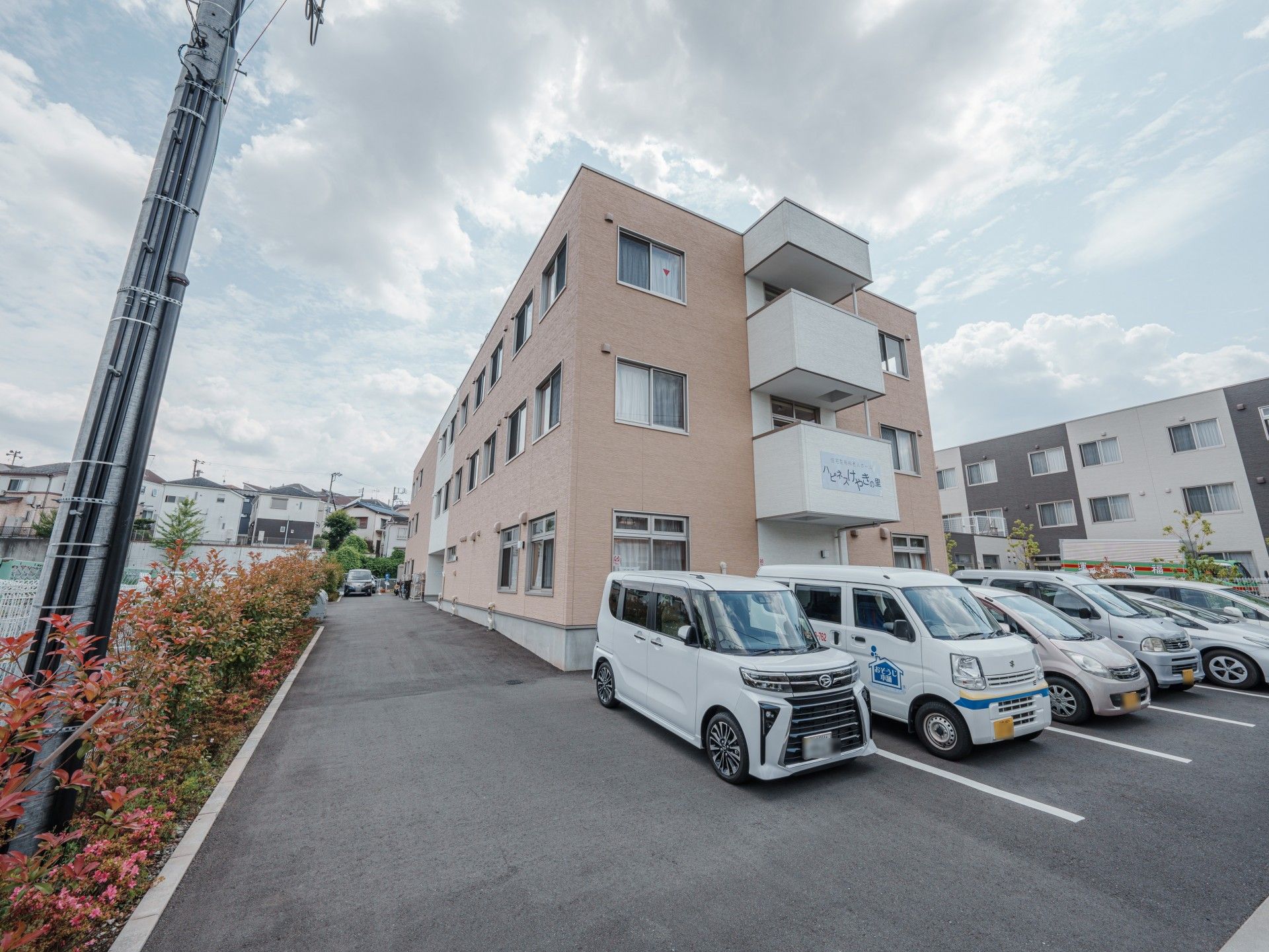 3階建ての建物はブラウン系の外観で角フロアにベランダがあり、駐車場には車が止まっている。