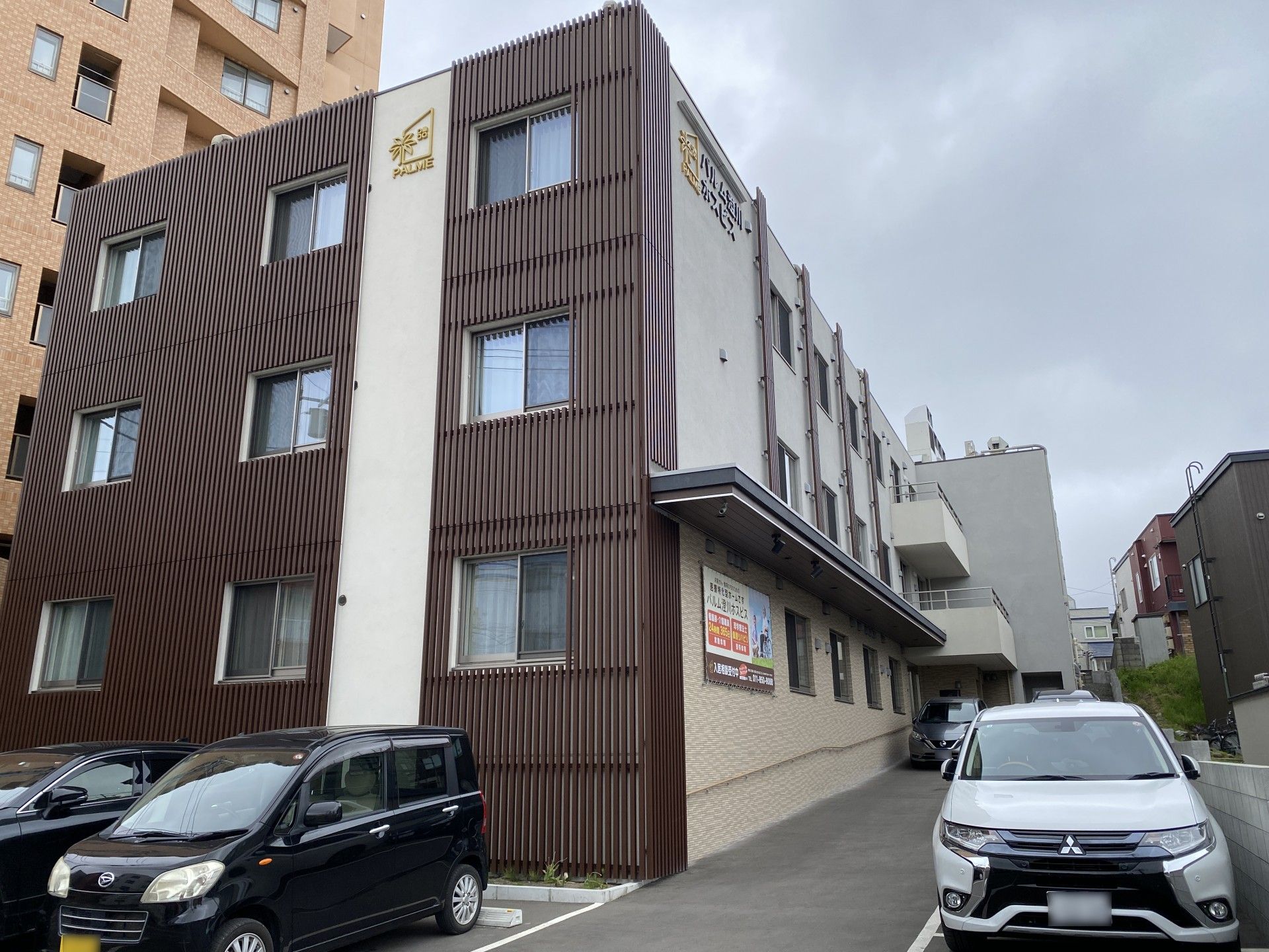 3階建ての建物の2階と3階にはベランダがあり、玄関には屋根が付き、白とブラウンの外壁。