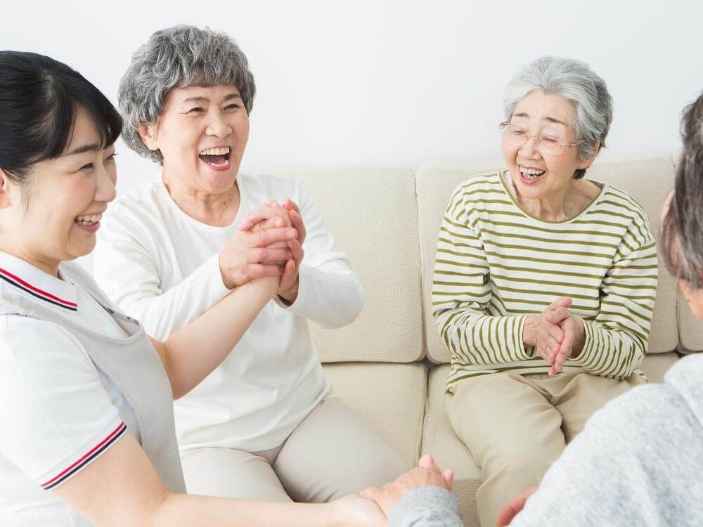 ベージュのソファを囲んで楽しそうに話している高齢女性たちと笑顔で過ごす白衣を着た女性スタッフ。