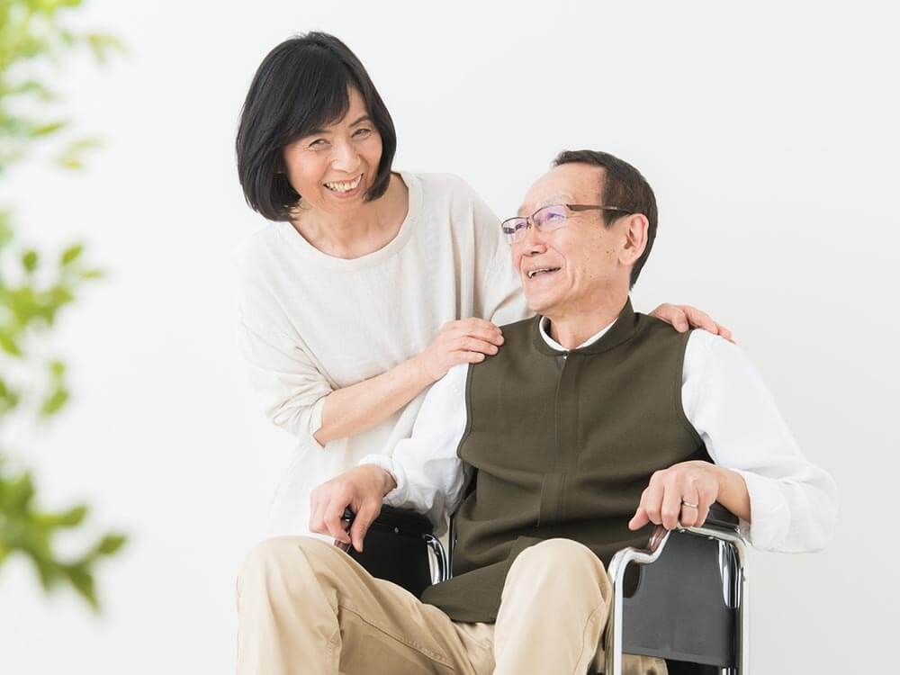 車いすの高齢男性のそばには白いゆったりとした服を着た高齢女性が立ち、笑顔で話している。