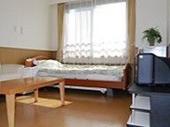 レースカーテンとカーテンが付いた窓がある部屋には寝具付きのベッドやテレビなどが置かれている。