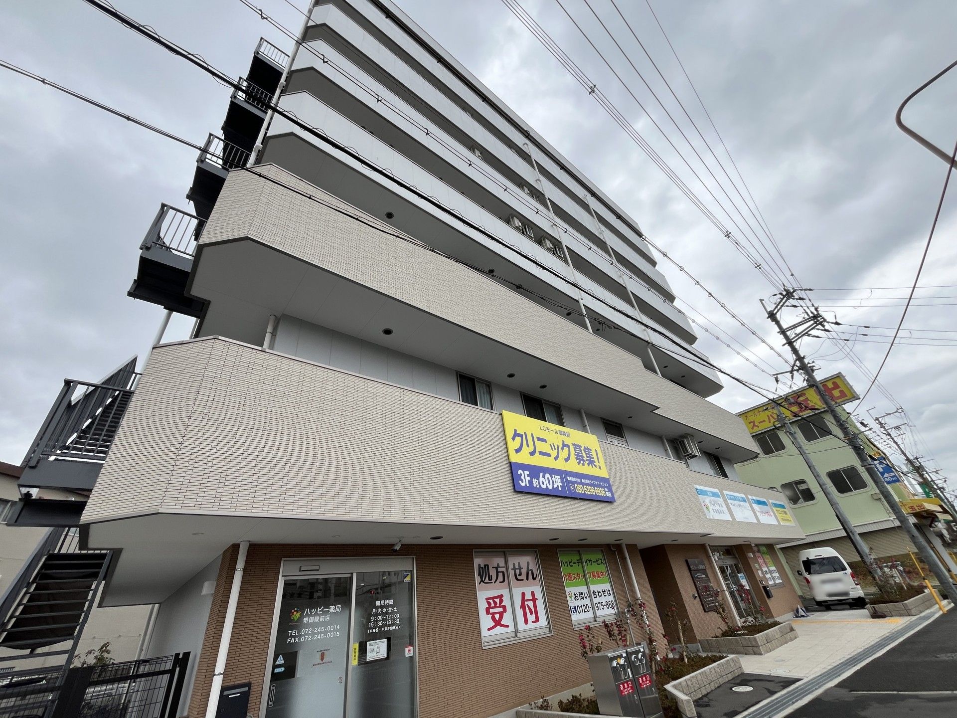 周辺には民家などがある場所に建っている背の高いベージュの外壁の建物にはベランダと屋外階段がある。