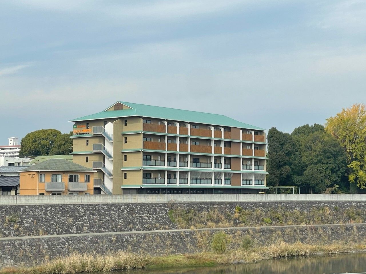 グリーンの屋根が付いた5階建ての建物はベージュの外壁で、周辺には緑や住宅がある。