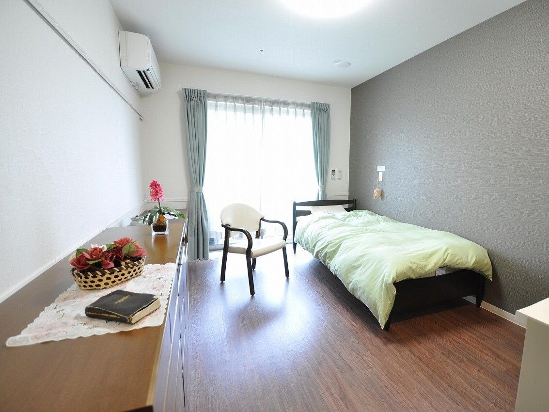 寝具付きのベッドが置かれたフローリングの部屋にはタンスなどもあり、ベッドサイドには緊急通報装置がある。