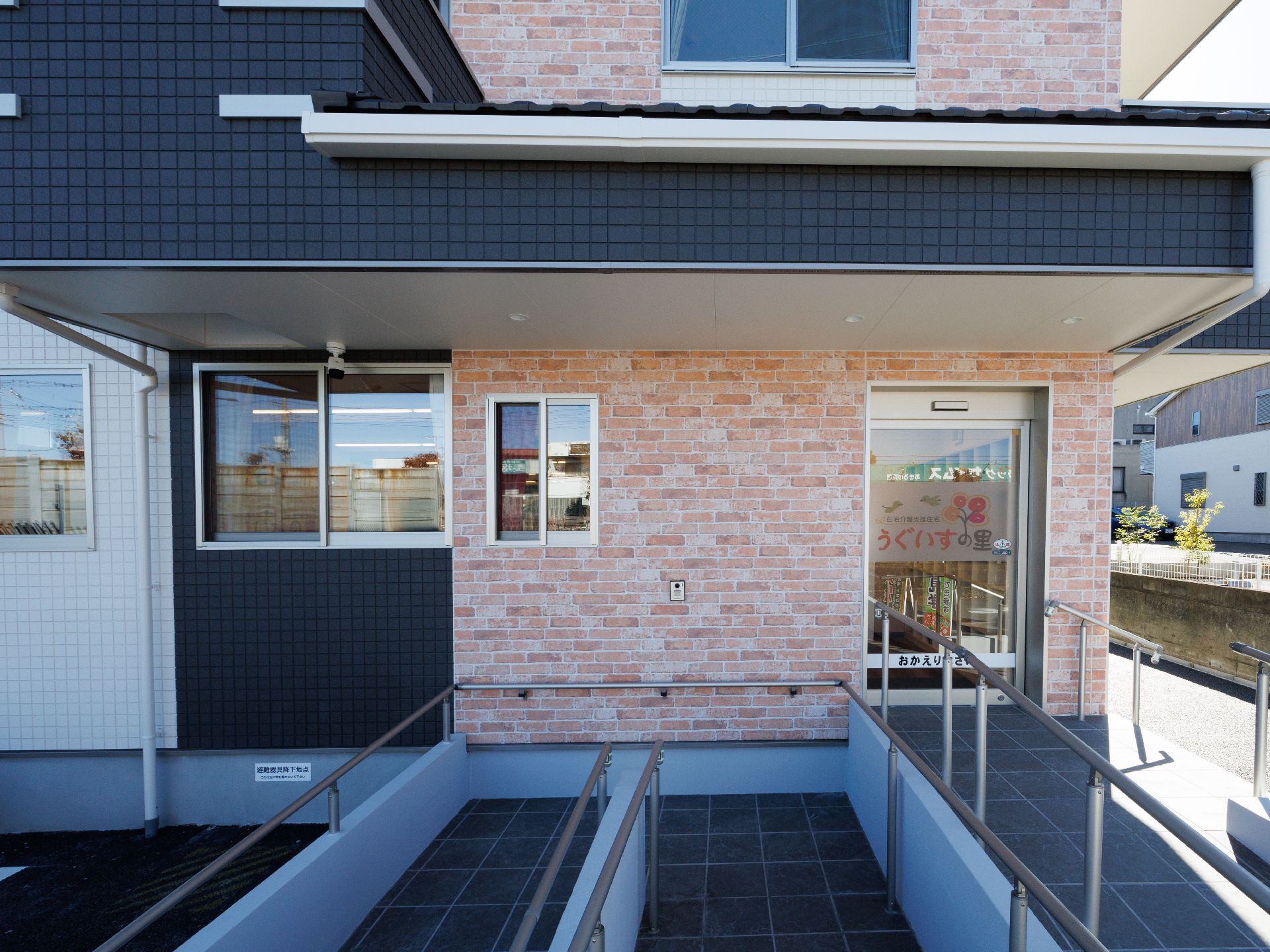 3階建ての建物は白を基調とした外壁で、各フロアにベランダがあり、玄関先には手すり付きのスロープがある。