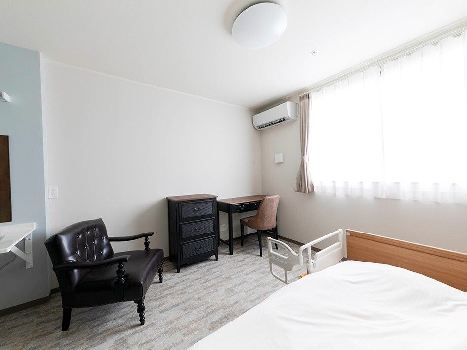 レースカーテンとカーテンが付いた大きな窓がある部屋には介護ベッドや家具が置かれている。