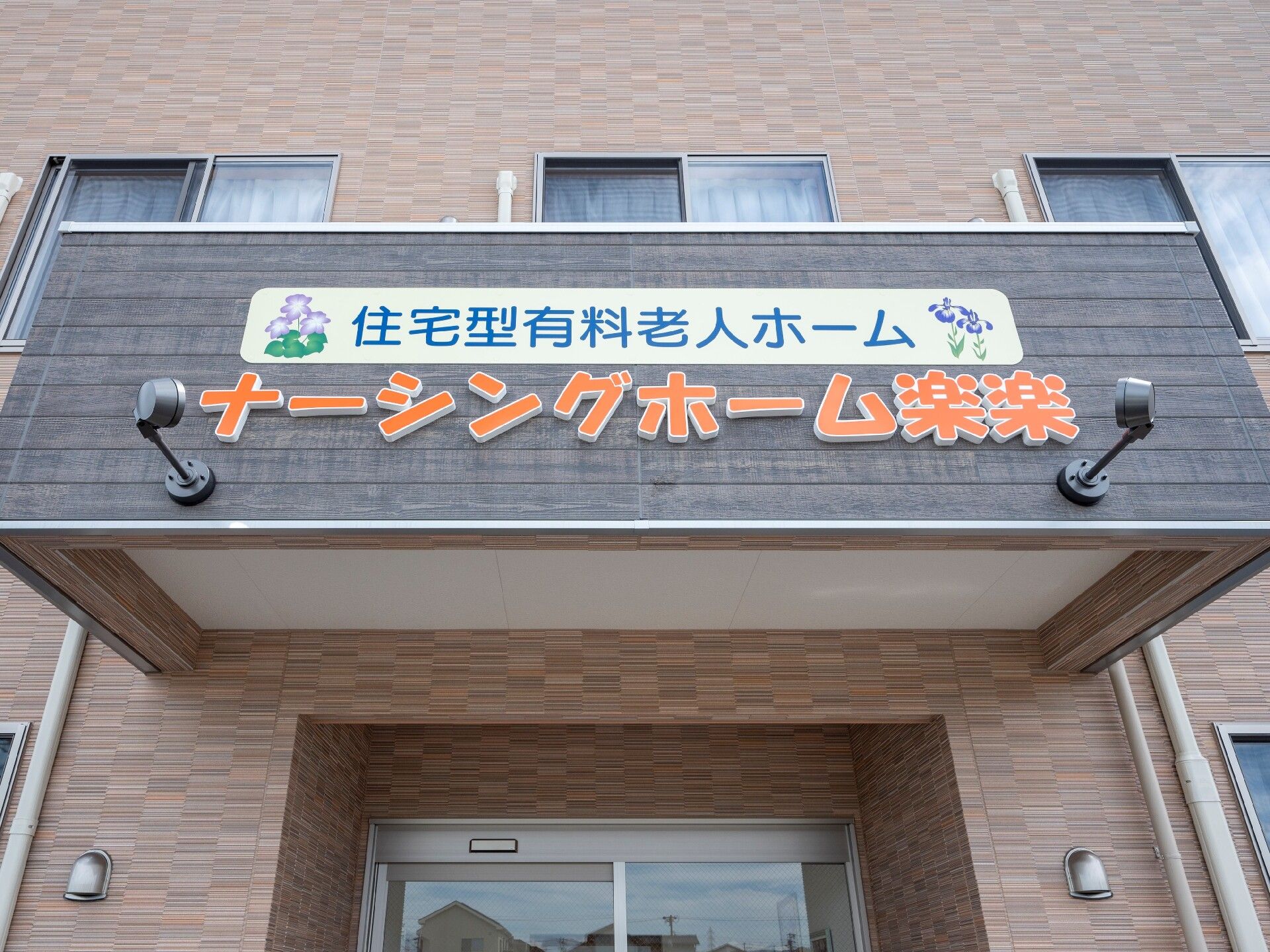 ブラウン系の建物は横に長く、シンプルな設計。2階建てで、そばには緑もある。